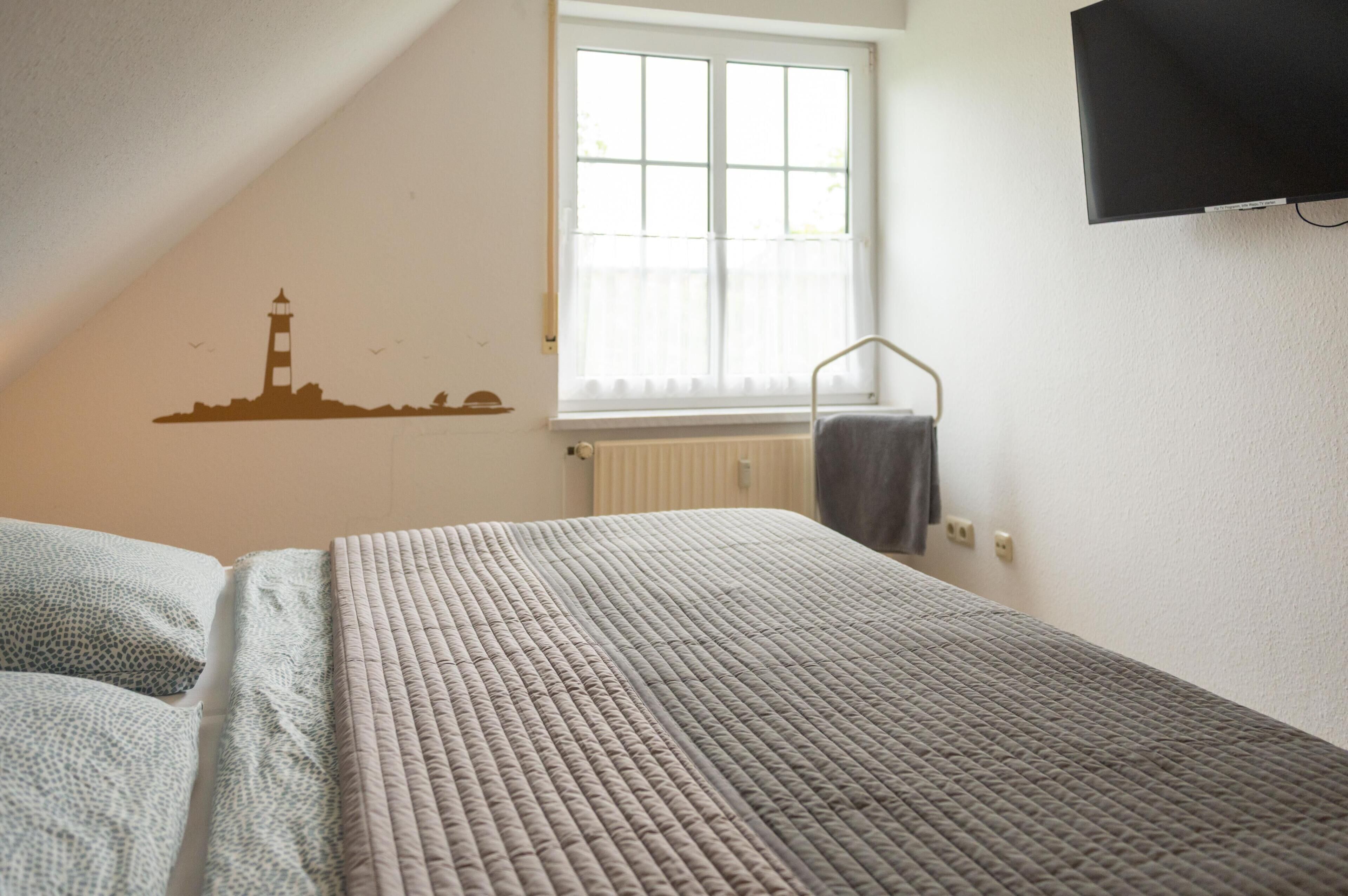 A bedroom with a bed, a window with blinds, a wall-mounted TV, and a radiator.