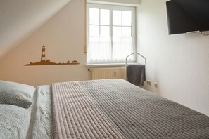 A bedroom with a bed, a window with blinds, a wall-mounted TV, and a radiator. - Stylish vacation apartment with balcony, modern furnishings and central location in Hooksiel - the Nordseeperle invites you to enjoy a relaxing vacation by the North Sea. (Hooksiel)