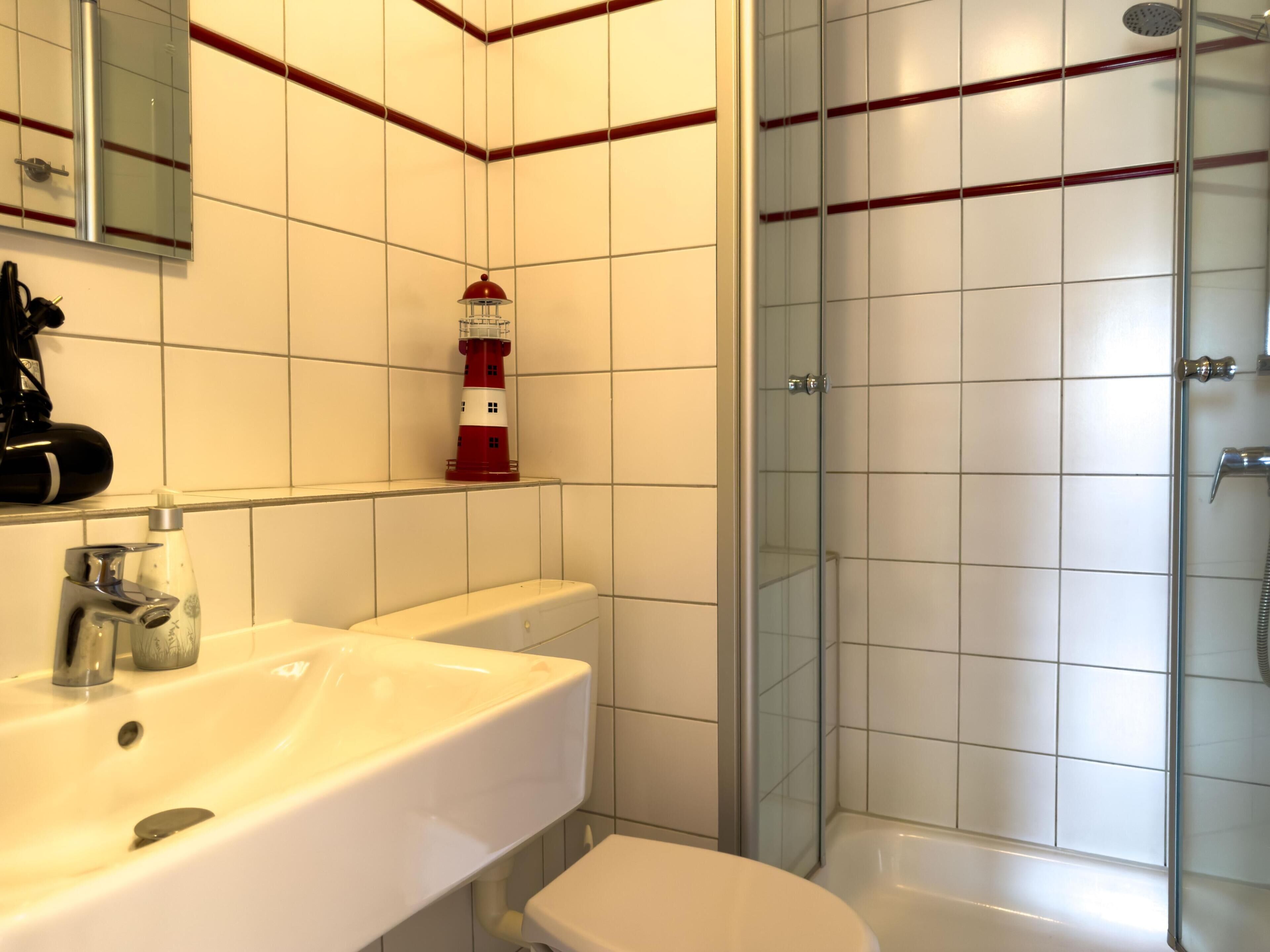Un baño con un lavamanos blanco, un inodoro, una ducha con cerramiento de vidrio y una decoración de faro rojo y blanco en la pared.