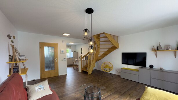 Een woonkamer met een rode bank, gele kussens, een glazen salontafel, een houten trap, een televisie en een keukenhoek.