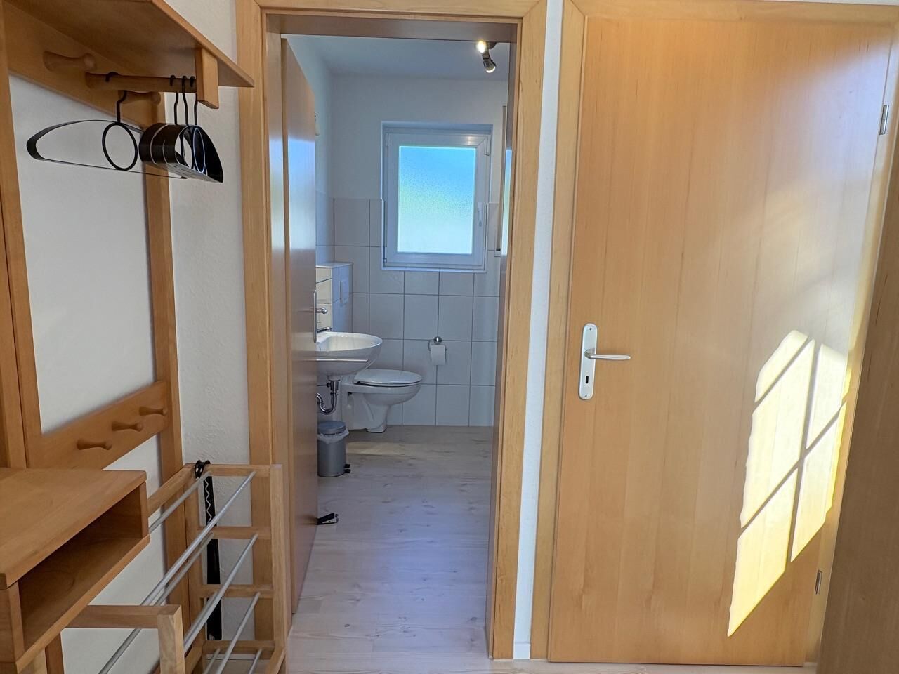 Un couloir étroit mène à une salle de bain équipée d’une fenêtre, d’un lavabo et de toilettes.