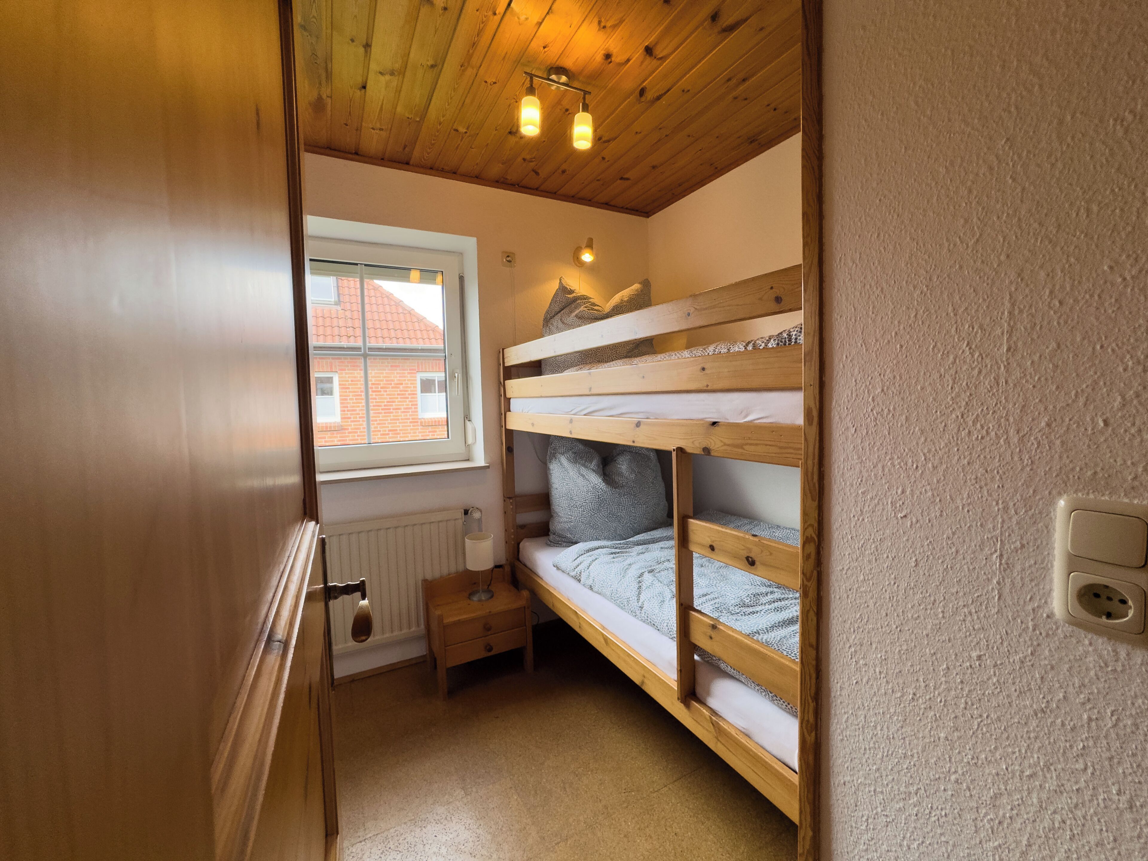 Een kleine, knusse kamer met een houten stapelbed, een raam met uitzicht op een bakstenen gebouw, een radiator en een klein houten nachtkastje.