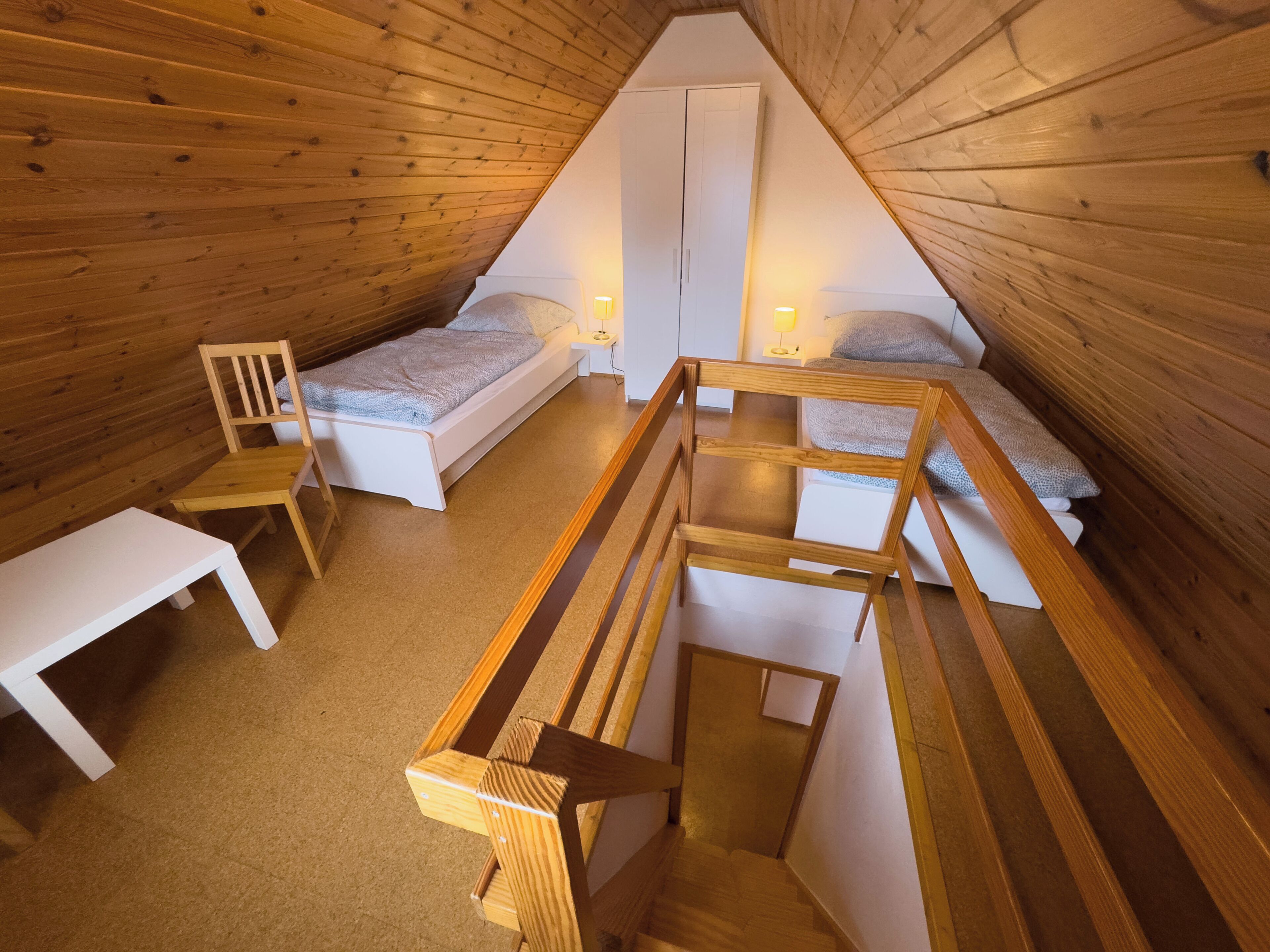 A triangular attic bedroom with two single beds, a wooden chair, a small table, and a white wardrobe.