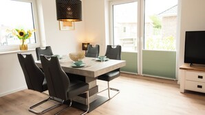 A dining area with a table, chairs, a vase with sunflowers, a hanging lamp, and a TV on a stand. - The modern vacation home Wanger Meerblick offers high-quality living comfort, three bedrooms, a fenced garden and a wonderful view of the Wangermeer - perfect for families and dog owners. (Wangerland)