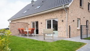 A brick house with a patio, a barbecue grill, and a dining area with chairs. - The modern vacation home Wanger Meerblick offers high-quality living comfort, three bedrooms, a fenced garden and a wonderful view of the Wangermeer - perfect for families and dog owners. (Wangerland)