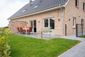 A brick house with a patio, a barbecue grill, and a dining area with chairs. - The modern vacation home Wanger Meerblick offers high-quality living comfort, three bedrooms, a fenced garden and a wonderful view of the Wangermeer - perfect for families and dog owners. (Wangerland)
