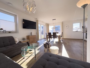 A modern living room with a sofa, glass coffee table, and a TV mounted on the wall. - The Wangermeer Perle I is a modern, spacious semi-detached house on the Wangermeer - ideal for families and groups looking for comfort and proximity to nature. (Wangerland)