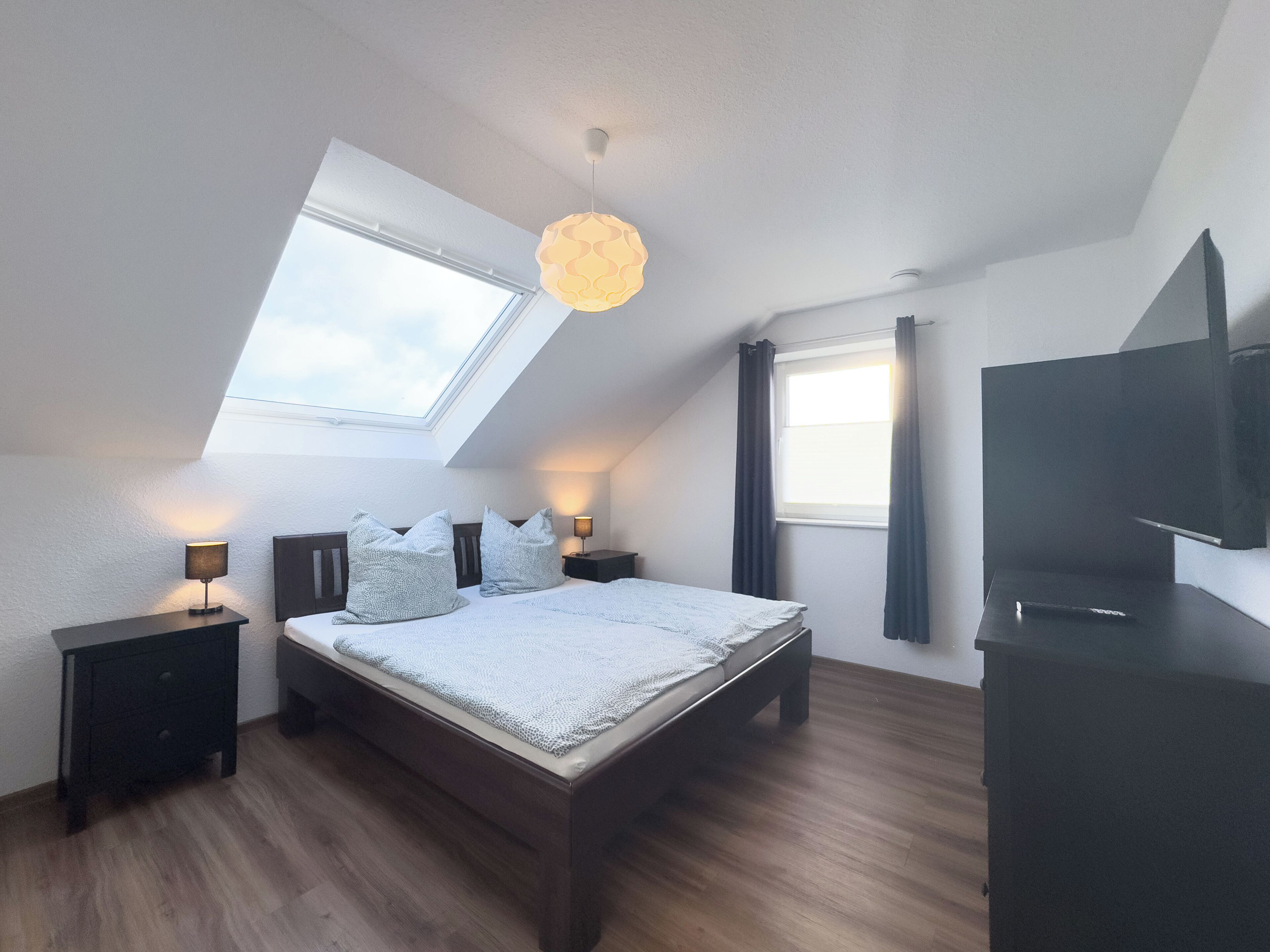 A bedroom with a skylight, a bed with pillows, a bedside table with a lamp, a TV, and a dark wardrobe.