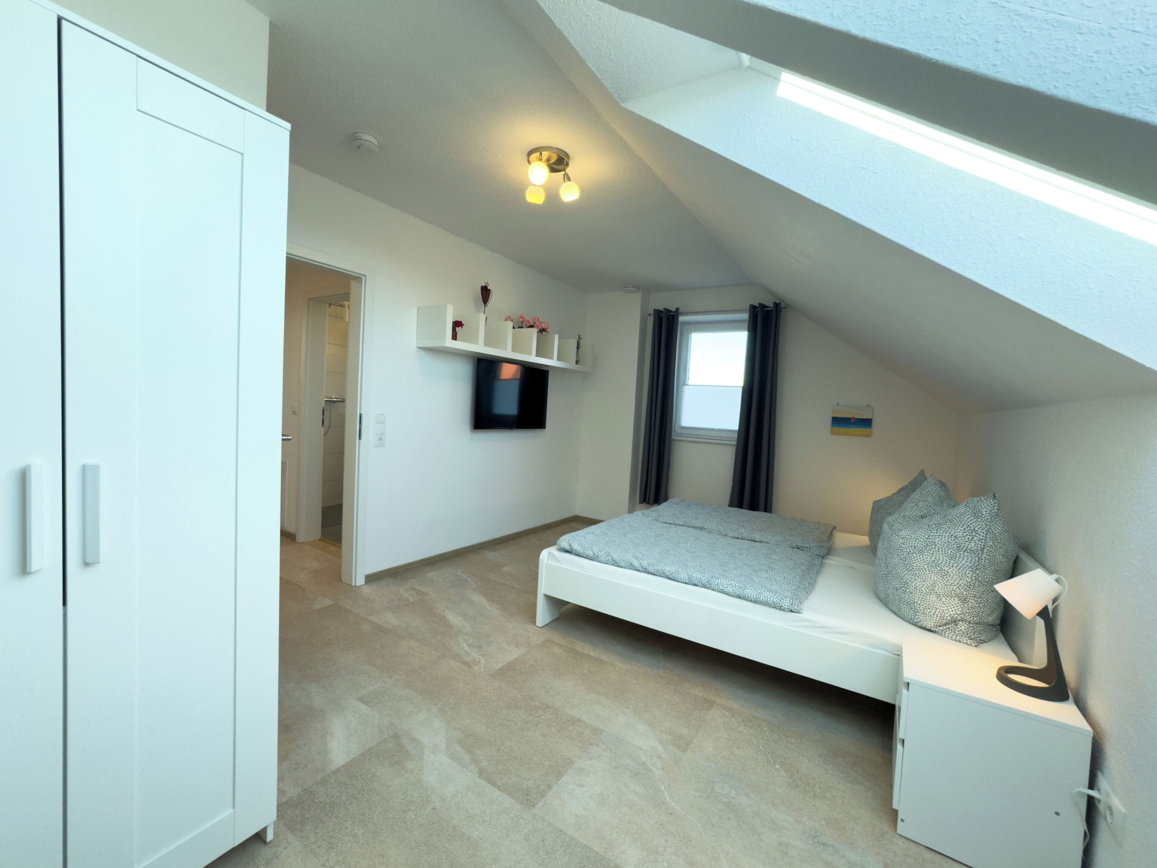 A bedroom with a bed, a TV, a shelf with flowers, a window, and a door.