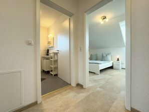 A bedroom with a bed, bedside table, and a nightstand. There is a bathroom with a sink and shelves. The room has a carpeted floor and white walls. - The Wangermeer Perle II is a modern semi-detached house on the Wangermeer and offers a cozy retreat for couples and families. (Wangerland)