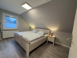 A bedroom with a bed, bedside tables, a window, and a ceiling light. - The Windfang 1 vacation home in Hooksiel offers modern comfort, a quiet location and a sunny terrace - perfect for relaxing days by the North Sea. (Hooksiel)
