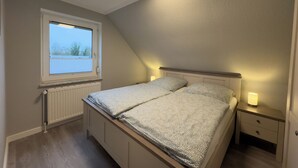 A bedroom with a bed, a nightstand, a window, and a ceiling light. - The Windfang 2 vacation home in Hooksiel offers modern comfort, a quiet location and a sunny terrace - perfect for relaxing days by the North Sea. (Hooksiel)