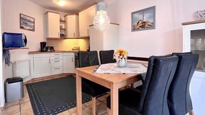 Een compacte keuken met witte kastjes, een eettafel met zwarte stoelen, een hanglamp en een kleine prullenbak.