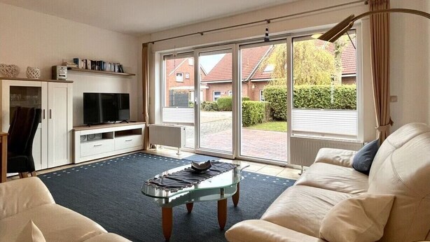Een woonkamer met een bank, een glazen salontafel, een tv op een dressoir en een grote schuifpui naar een tuin.