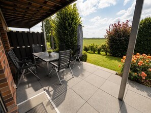 An outdoor patio with a table and chairs, surrounded by a wooden fence and a view of a garden with flowers and a hedge. - Comfortable vacation home "Austernfischer" in Wangerland near the North Sea with sauna, 3 bedrooms for up to 5 guests - dogs welcome. (Wangerland)