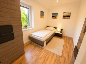 A bedroom with a bed, bedside table, lamp, and a window with a view of greenery. - Comfortable vacation home "Austernfischer" in Wangerland near the North Sea with sauna, 3 bedrooms for up to 5 guests - dogs welcome. (Wangerland)