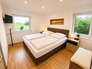 A modern bedroom with a large bed, bedside tables, a TV, and a view of greenery. - Comfortable vacation home "Austernfischer" in Wangerland near the North Sea with sauna, 3 bedrooms for up to 5 guests - dogs welcome. (Wangerland)