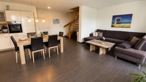 Una sala moderna con un sofá marrón oscuro, un comedor con mesa y sillas de madera, y una cocina con armarios blancos y un horno empotrado.