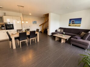 A modern living room with a dark brown sofa, a dining area with a wooden table and chairs, and a kitchen with white cabinets and a built-in oven. - Comfortable vacation home "Austernfischer" in Wangerland near the North Sea with sauna, 3 bedrooms for up to 5 guests - dogs welcome. (Wangerland)