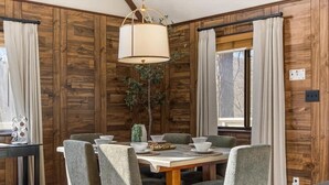 Un comedor con pared de paneles de madera, una mesa dispuesta para seis personas con sillas, una lámpara colgante y una ventana con cortinas.
