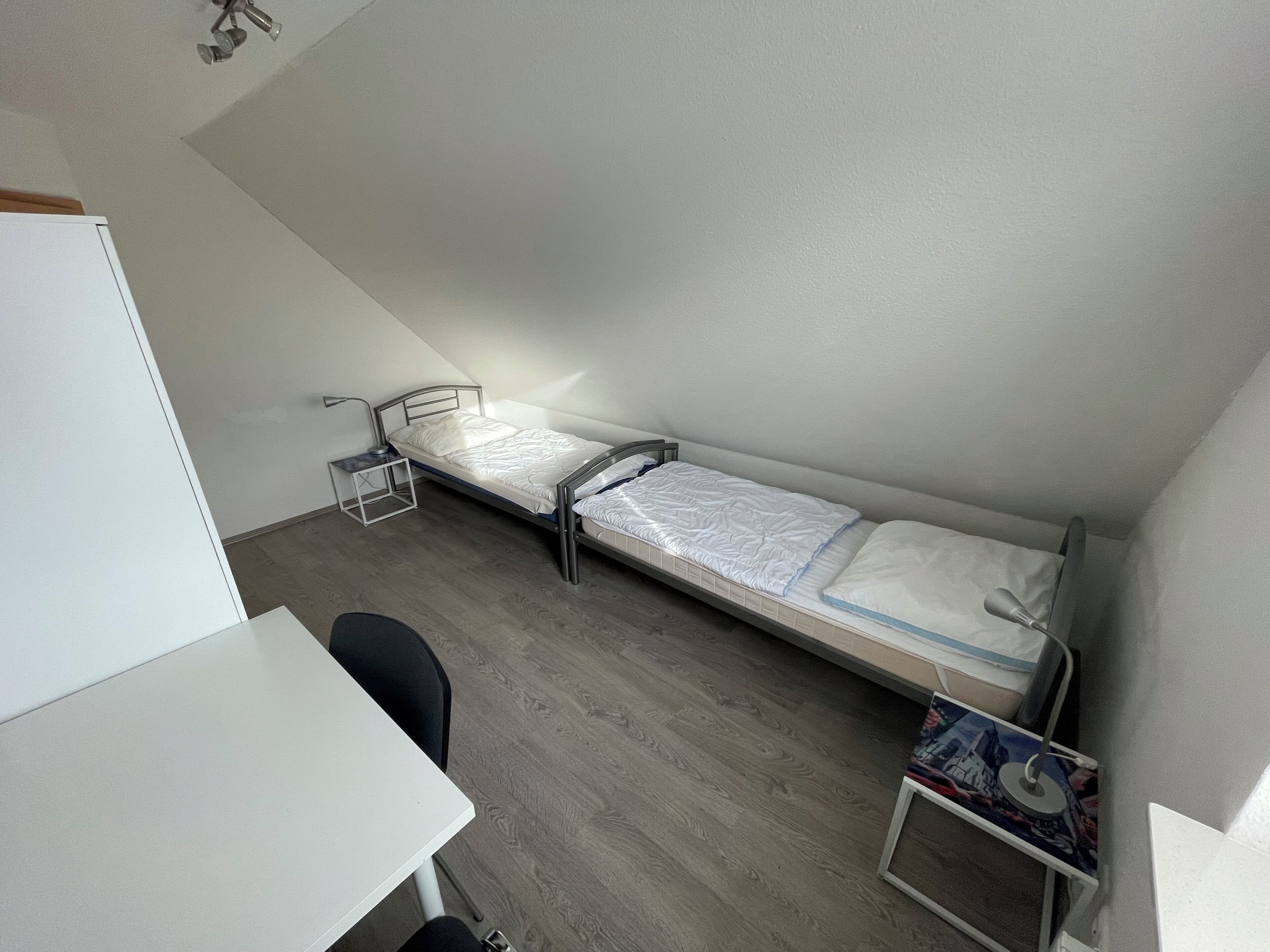 Una habitación pequeña con cama individual, escritorio, silla, mesita de noche y un estante.