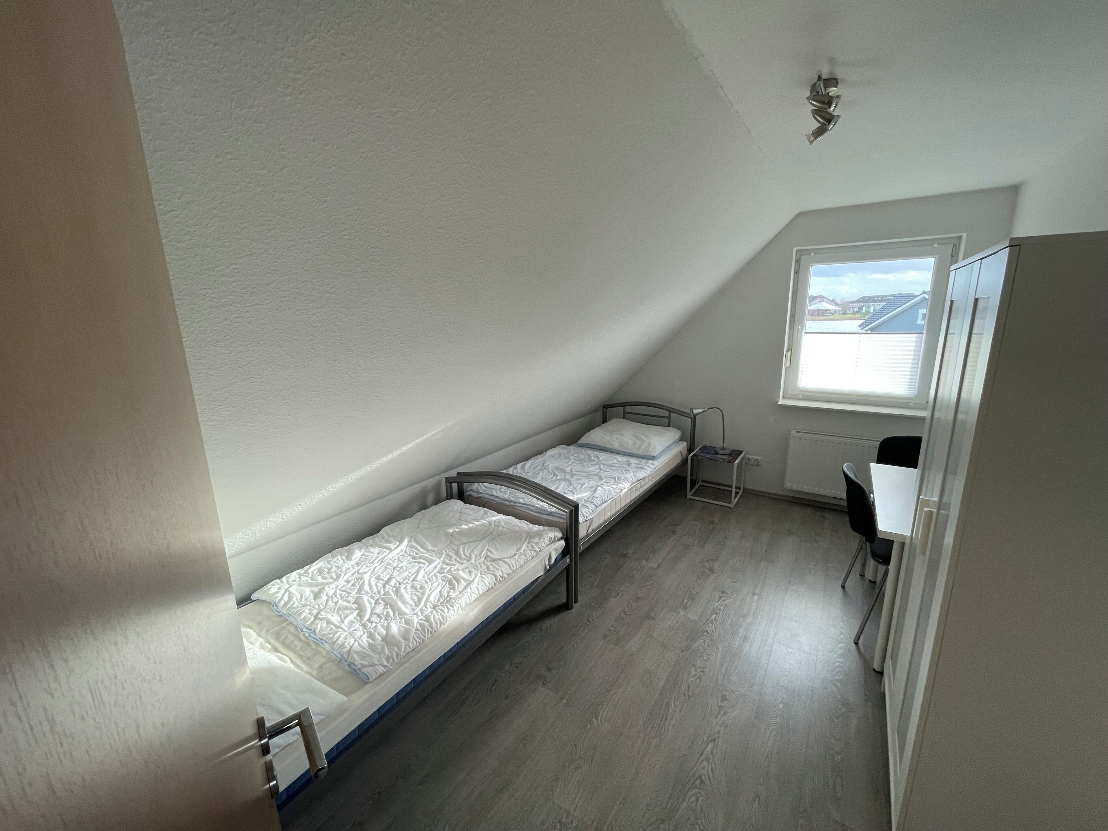 Um apartamento pequeno, de um único quarto, com teto inclinado, uma janela, uma escrivaninha e uma cama.