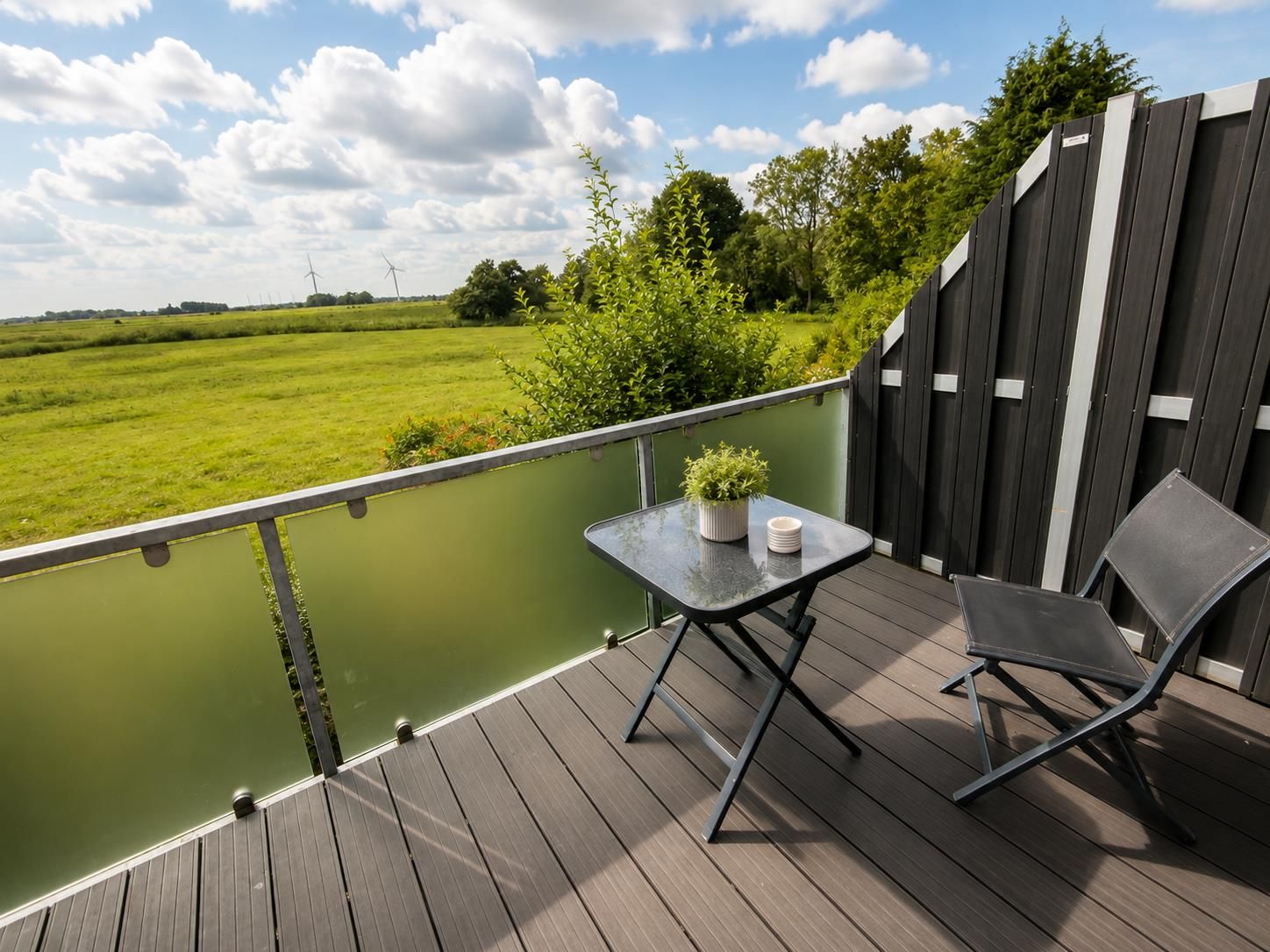 A balcony with a table and chairs, overlooking a green landscape with trees and wind turbines.