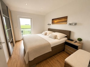 A modern bedroom with a large bed, a bedside table, a small bedside plant, and a view of the outdoors through a glass door. - Cozy vacation home "Sturmmöwe" in Wangerland near the North Sea with sauna, 2 bedrooms for up to 3 guests - dogs welcome. (Wangerland)