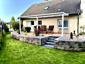 A house with a patio, a lawn, and a stone retaining wall. - "Rosenwind" Vacation Home in Wangerland: 5 bedrooms for up to 9 people, 2 bathrooms, wood-burning stoves, a large kitchen, and a garden with a porch—perfect for a vacation on the North Sea. (Wangerland)