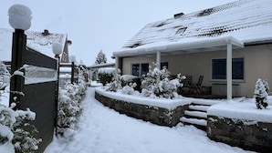 Zona residencial plena de neu, amb una casa, un porxo cobert i una paret de pedra.