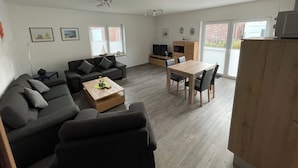 Ein modernes Wohnzimmer mit Sofa, Couchtisch und Esstisch. Es gibt Deckenventilatoren, einen Fernseher und ein Fenster mit Jalousien.