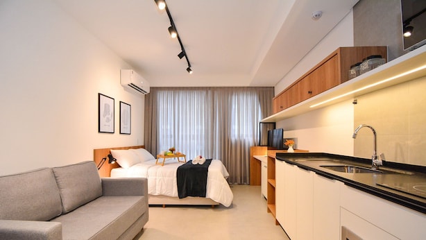 A compact hotel room with a bed, a sofa, a kitchenette, and a large window with curtains. - Studio em frente a UFSC e HU - 319A (Florianópolis)