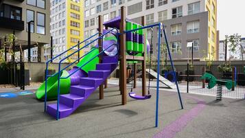 Une aire de jeux avec un toboggan violet, une structure d’escalade aux couleurs verte et violette, et des balançoires.