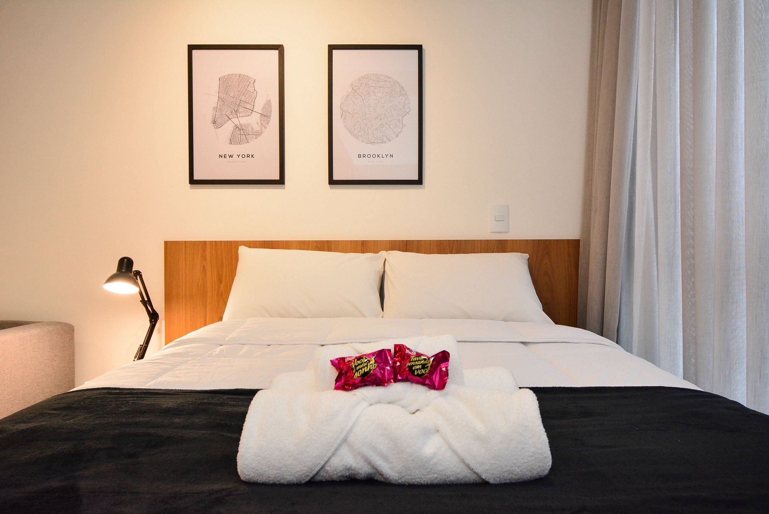 Habitación de hotel con cama, mesita de noche con lámpara, dos mapas enmarcados en la pared y una toalla con aperitivos sobre la cama.