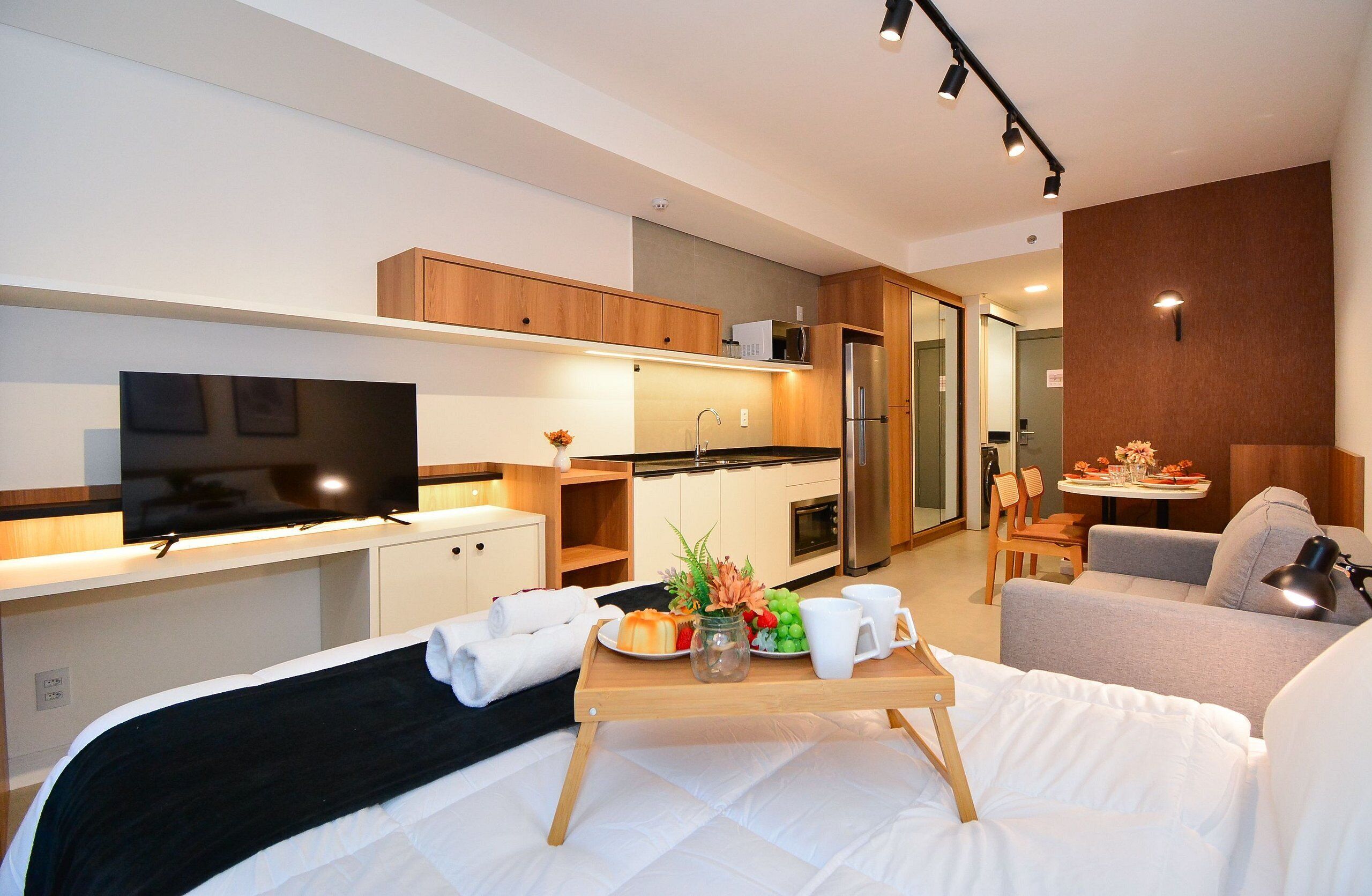 A modern hotel room with a bed, a TV, a kitchenette, and a dining area.