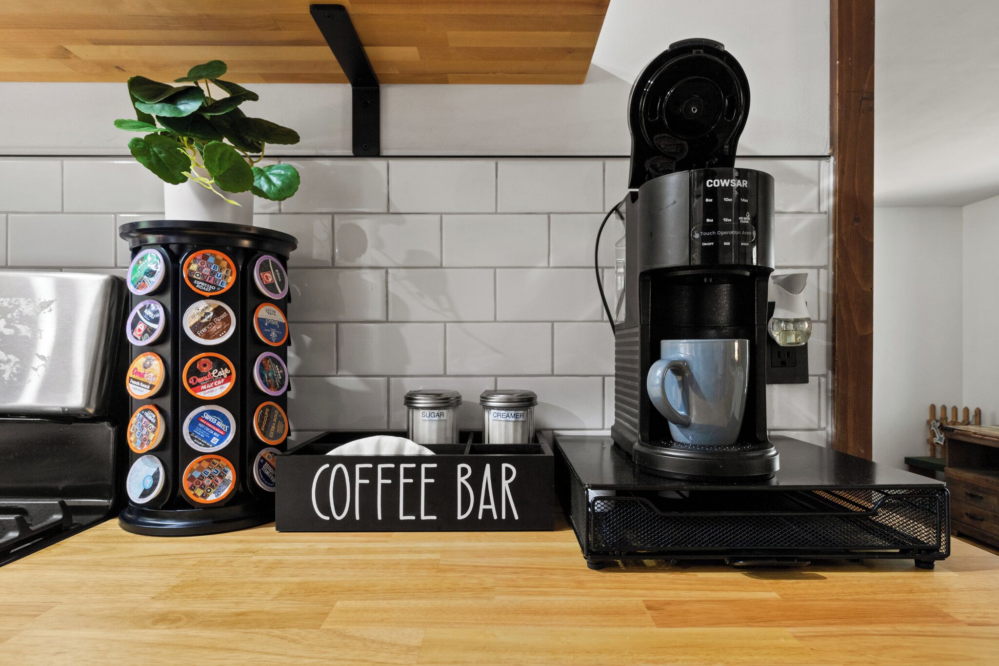 Un espace café aménagé avec une machine à café, un récipient contenant des dosettes de café, des sucriers et des pots de crème, ainsi qu’une enseigne indiquant « CAFÉ ».