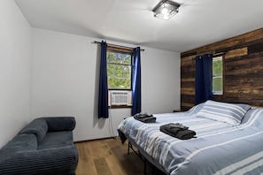 A bedroom with a bed, a chair, a window with curtains, and a wooden wall. - Charming Cottage by Lake Ontario | Firepit + Yard (Richland)