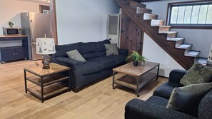 A living room with a sofa, two coffee tables, a lamp, a potted plant, a staircase, and a kitchen area with a microwave and refrigerator.