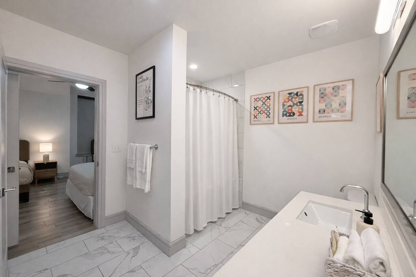 Une salle de bain moderne avec un grand miroir, une baignoire et un rideau de douche. Des œuvres d’art encadrées ornent le mur, et un porte-serviettes est muni de serviettes.
