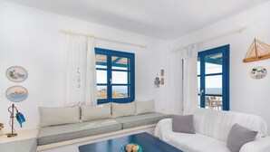 Een kamer met een bank, een bijzettafeltje en uitzicht door een raam met blauwe luiken.
