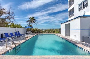 A swimming pool with blue chairs, a palm tree, and a white building with a blue roof. - Quiet north side condo. Beach access, inspiring views, pool. (Fort Myers Beach)