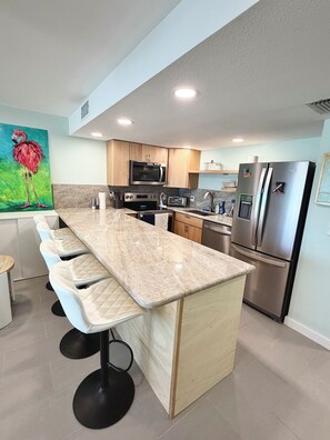 A modern kitchen with a central island, stainless steel appliances, and a large painting of a flamingo. - Quiet north side condo. Beach access, inspiring views, pool. (Fort Myers Beach)