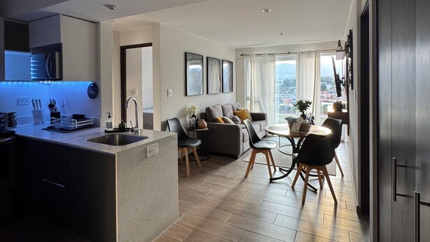 Een moderne keuken met een centraal eiland, een eethoek met tafel en stoelen, en een woonkamer met een bank en uitzicht op de stad.
