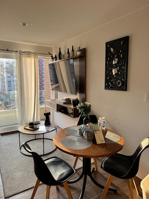 A modern dining area with a round table, black chairs, a TV mounted on the wall, and a balcony with a view of buildings.