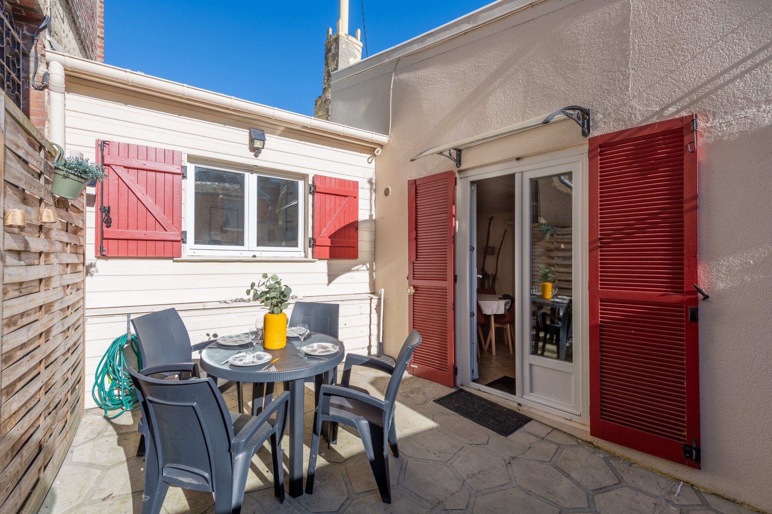 Un patio con una mesa redonda y sillas, una planta colgante y una puerta con persianas rojas.