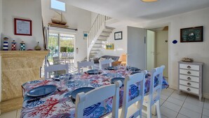 A dining area with a table set for six, white chairs, a staircase, and a view of the outdoors. - La Maison Orion - Oléron, à 1km de la plage (Dolus-d'Oléron)