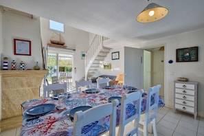 A dining area with a table set for six, white chairs, a staircase, and a view of the outdoors. - La Maison Orion - Oléron, à 1km de la plage (Dolus-d'Oléron)