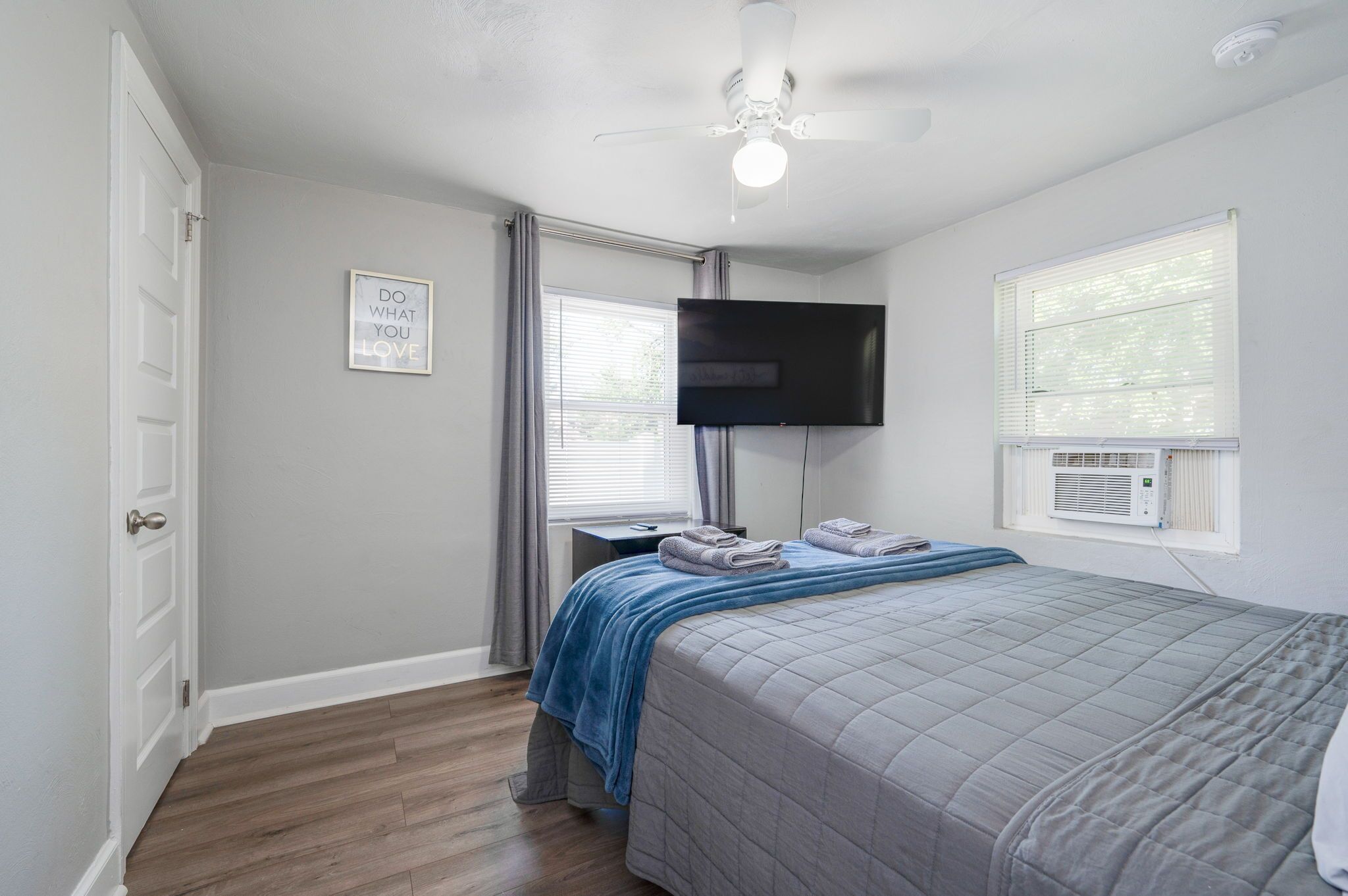 A bedroom with a bed, a flat-screen TV mounted on the wall, a ceiling fan, and a window with blinds.