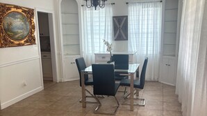 Un coin repas agrémenté d’un lustre, d’une horloge et offrant une vue sur l’extérieur à travers de fines rideaux.
