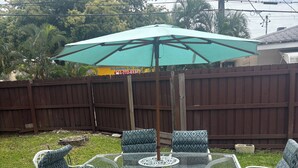 Une terrasse extérieure avec une table à plateau de verre et des chaises en osier blanc, entourée d’une clôture en bois et d’un parasol vert.
